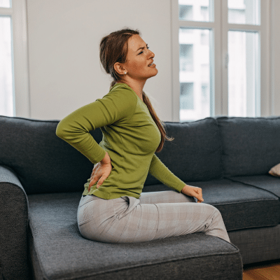 Common causes of back pain and when to see a specialist 3 Woman sitting on a couch, holding her lower back, dealing with back pain