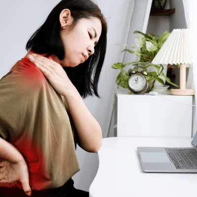 How poor posture affects spine health 5 Woman rubbing her shoulder and lower back in pain while sitting at a desk with a laptop, representing how poor posture affects your spine health
